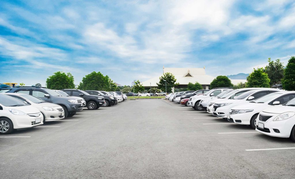Wide view of a large parking lot with rows of parked cars on both sides, leading toward a building in the distance under a partly cloudy sky.