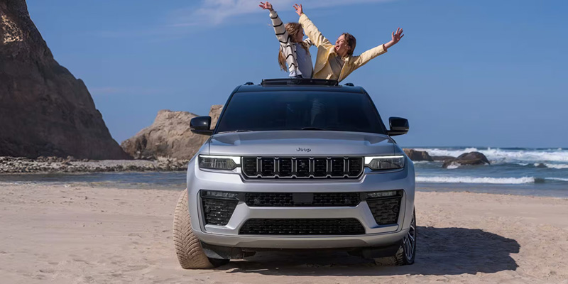 A silver Jeep Grand Cherokee parked on a beach with two people enjoying themselves, one standing on the roof and the other inside the vehicle, against a backdrop of ocean waves and cliffs.