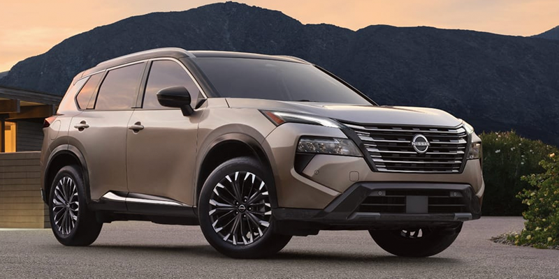 A beige Nissan Rogue parked in front of a house with mountains in the background during sunset.