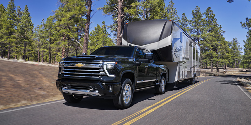 A black Chevrolet truck towing a large RV on a winding road surrounded by pine trees.