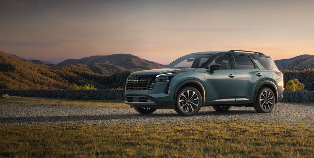 A teal Nissan SUV parked on gravel with mountains in the background during sunset.