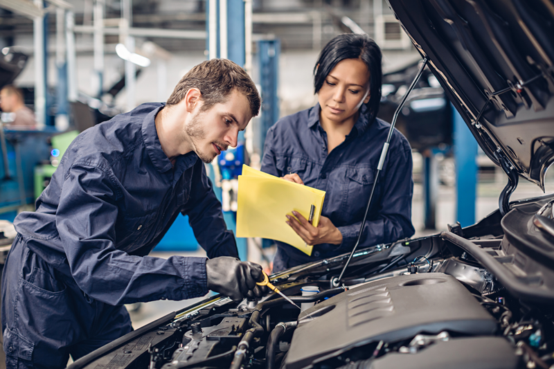 Two automotive technicians inspecting a vehicle engine and recording maintenance findings in a dealership service center.