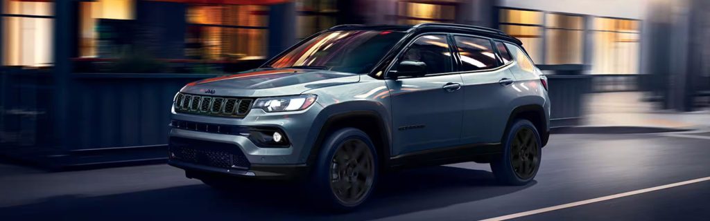 A blue SUV driving in an urban setting at dusk, with lights from nearby buildings illuminating the scene.