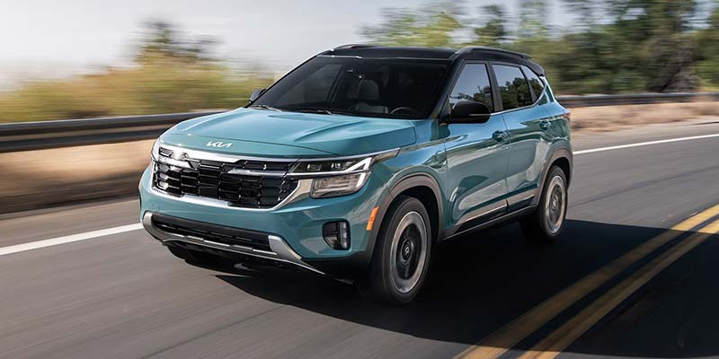A teal Kia Seltos SUV driving on a winding road surrounded by trees.
