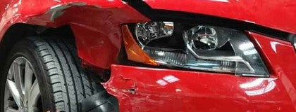Close-up of a damaged red car showing crumpled bodywork, a detached fender, and a partially obscured front tire.
