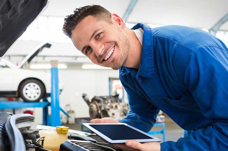A mechanic smiling while holding a tablet, working on a car in an automotive workshop.